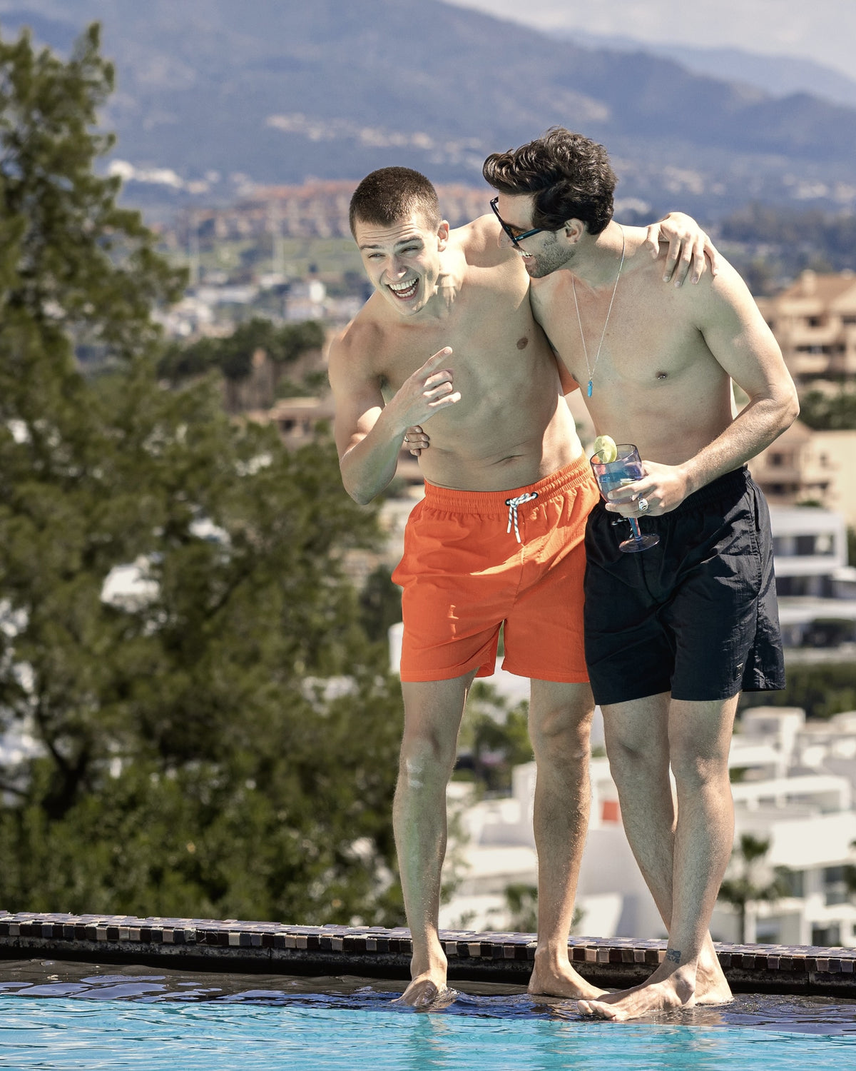 Zwei Männer am Poolrand, einer trägt rote Badehose 'Jay', der andere schwarze Badehose und hält ein Getränk