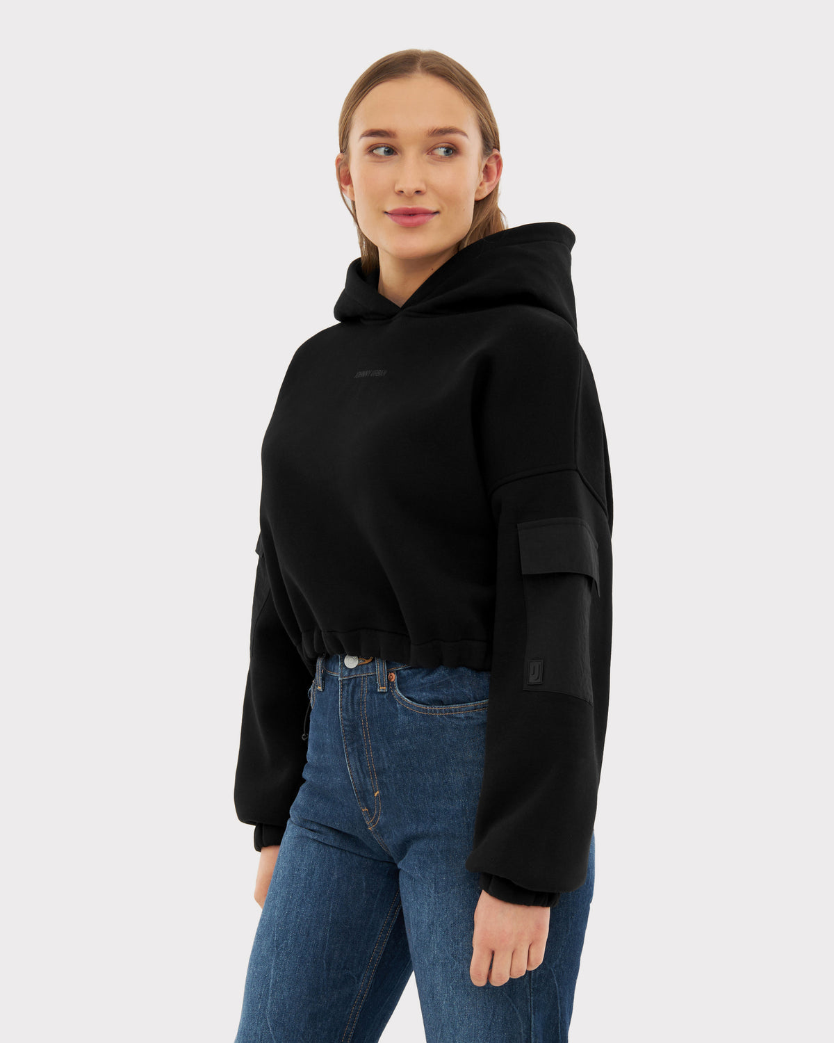 Frau trägt schwarzen Cropped Hoodie 'Carly' mit Kapuze und Tasche am Ärmel, kombiniert mit blauen Jeans