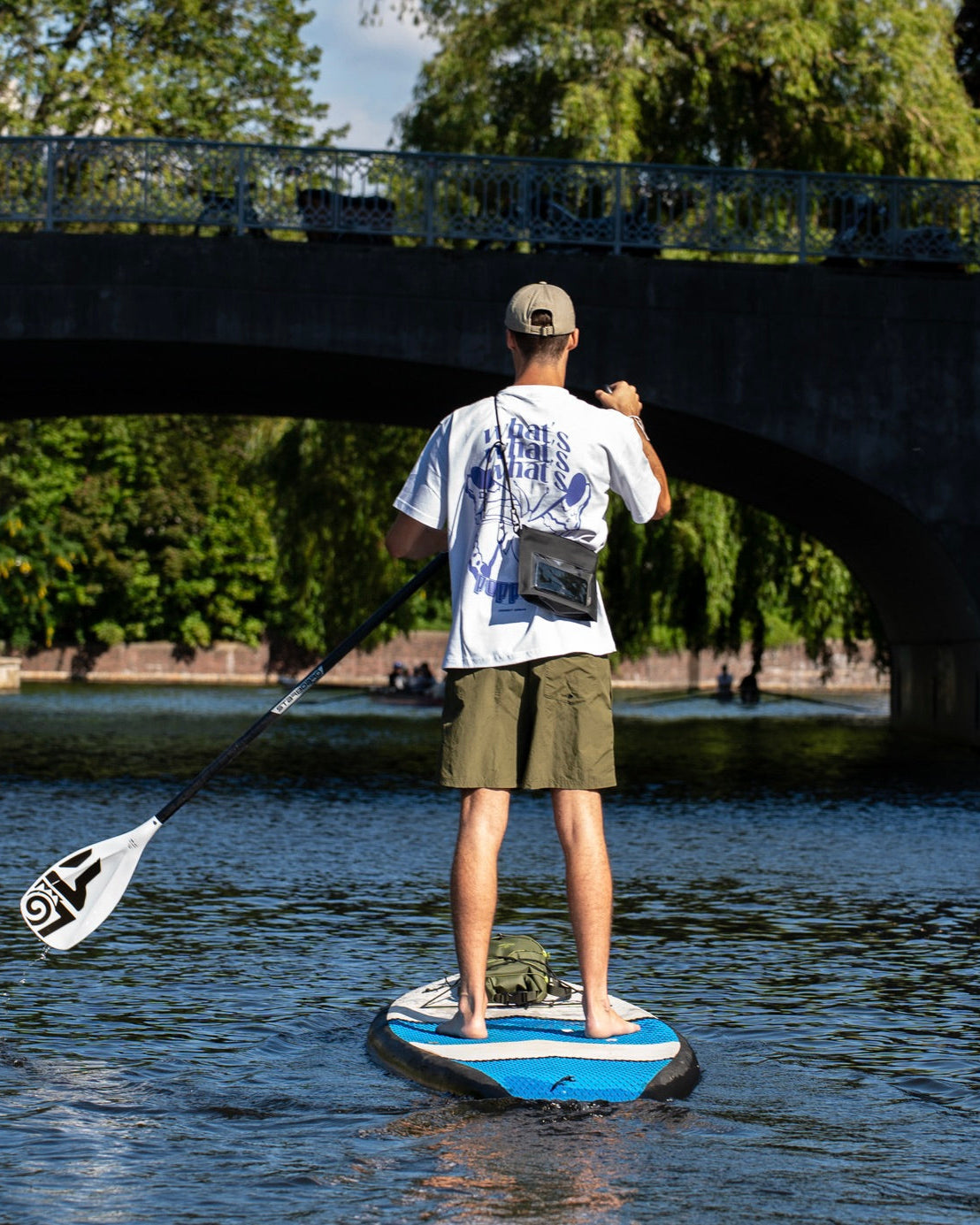 Rückansicht eines Mannes auf einem Stand-up-Paddle-Board mit Dry Bag 'Alfie' am Körper, umgeben von Wasser und Brücke im Hintergrund