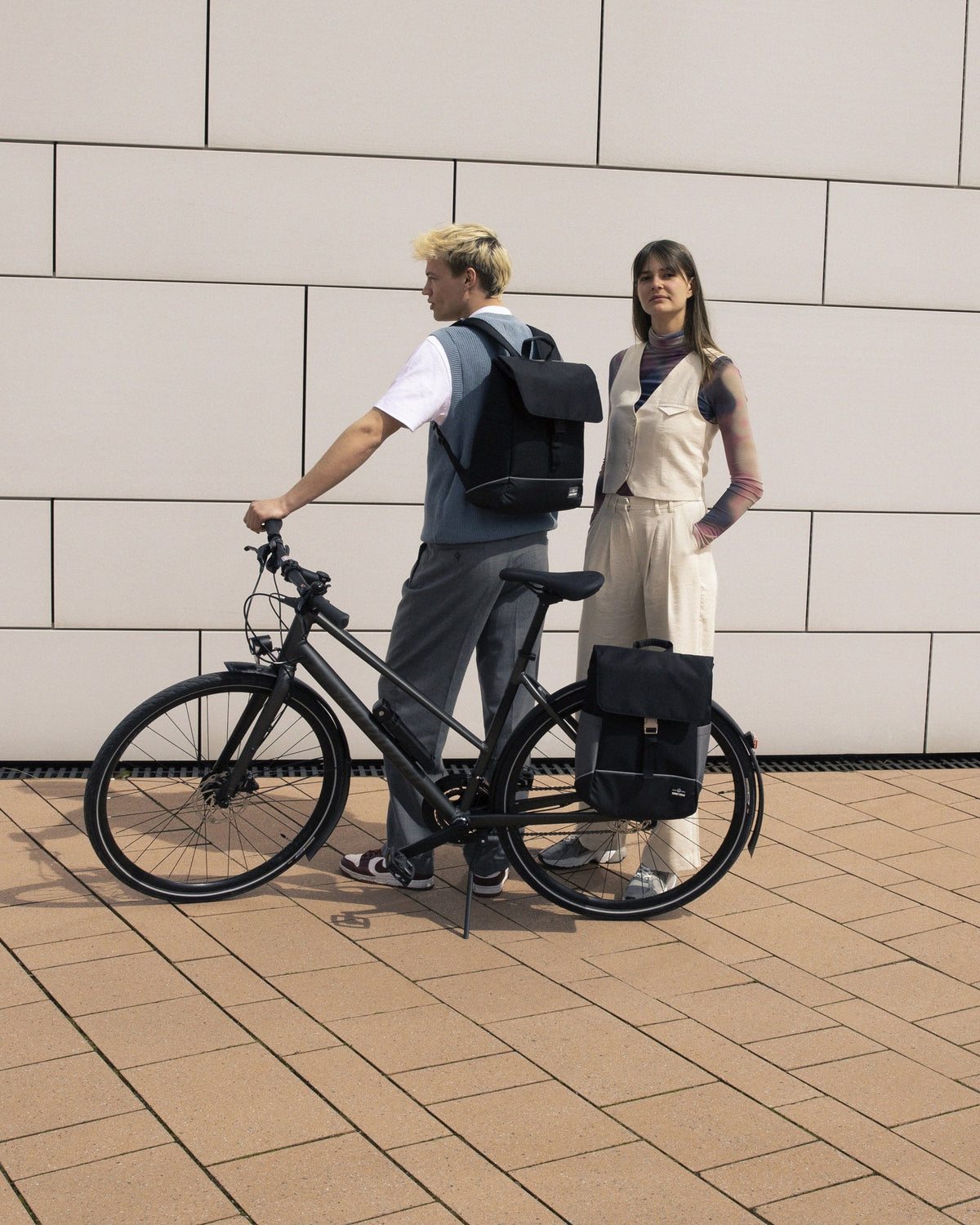Mann mit schwarzem Rucksack 'Mika' auf einem schwarzen Fahrrad, Frau neben ihm mit schwarzem Rucksack 'Mika' neben Fahrrad