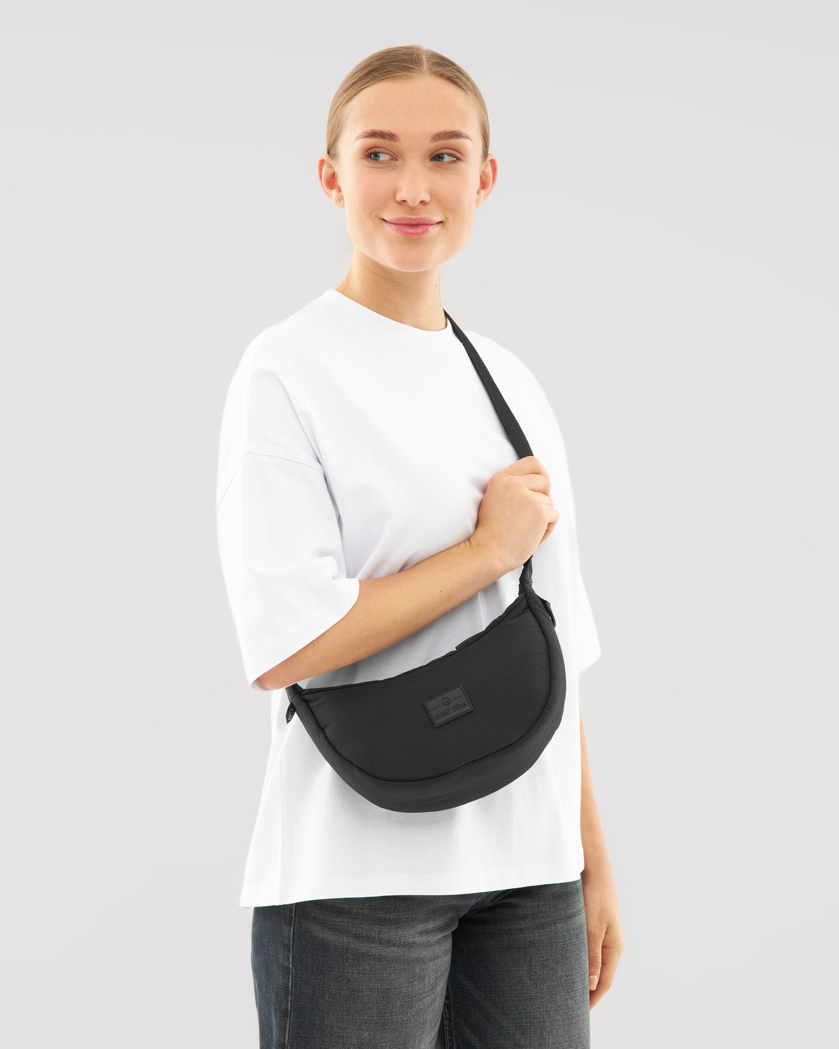 Frau trägt schwarze Half Moon Bag Kaia Small quer am Körper, weißes Oberteil, dunkle Jeans