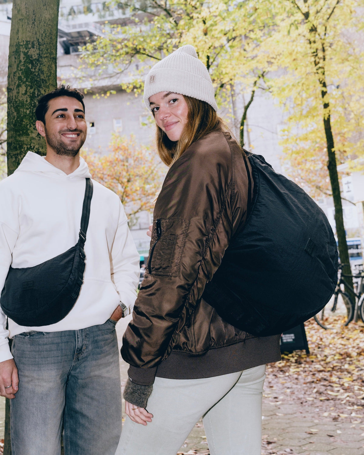 Mann mit hellbeigem Hoodie trägt schwarze Half Moon Bag Kaia XL crossbody, Frau mit brauner Jacke und weißer Mütze trägt sie als Rucksack