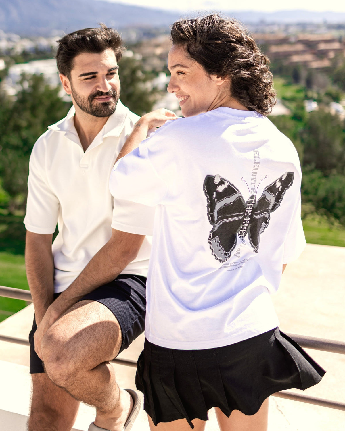 Frau trägt weißes Oversize-T-Shirt 'Sammy Chrome Butterfly' mit schwarzem Schmetterlingsmotiv auf dem Rücken, Mann daneben in weißem Poloshirt und schwarzen Shorts