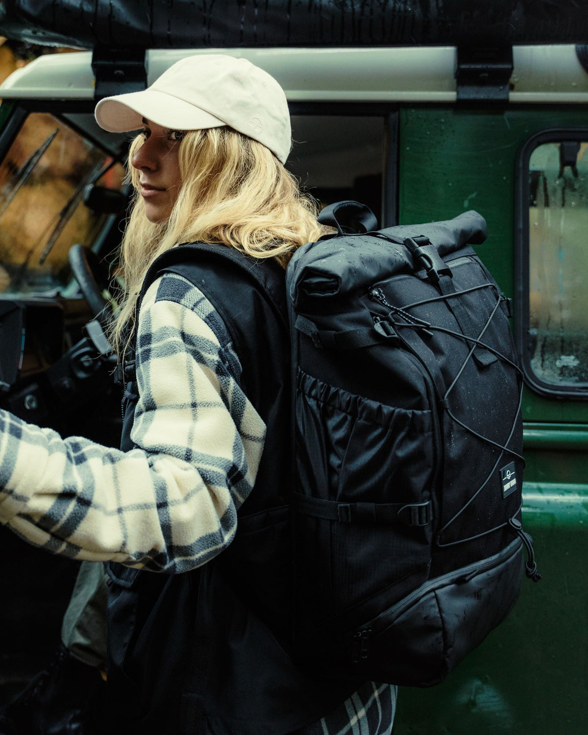 Frau mit hellbeiger Baseballkappe und kariertem Flanellhemd trägt schwarzen Reiserucksack 'Allen Large Travel' und öffnet grünes Fahrzeugtür
