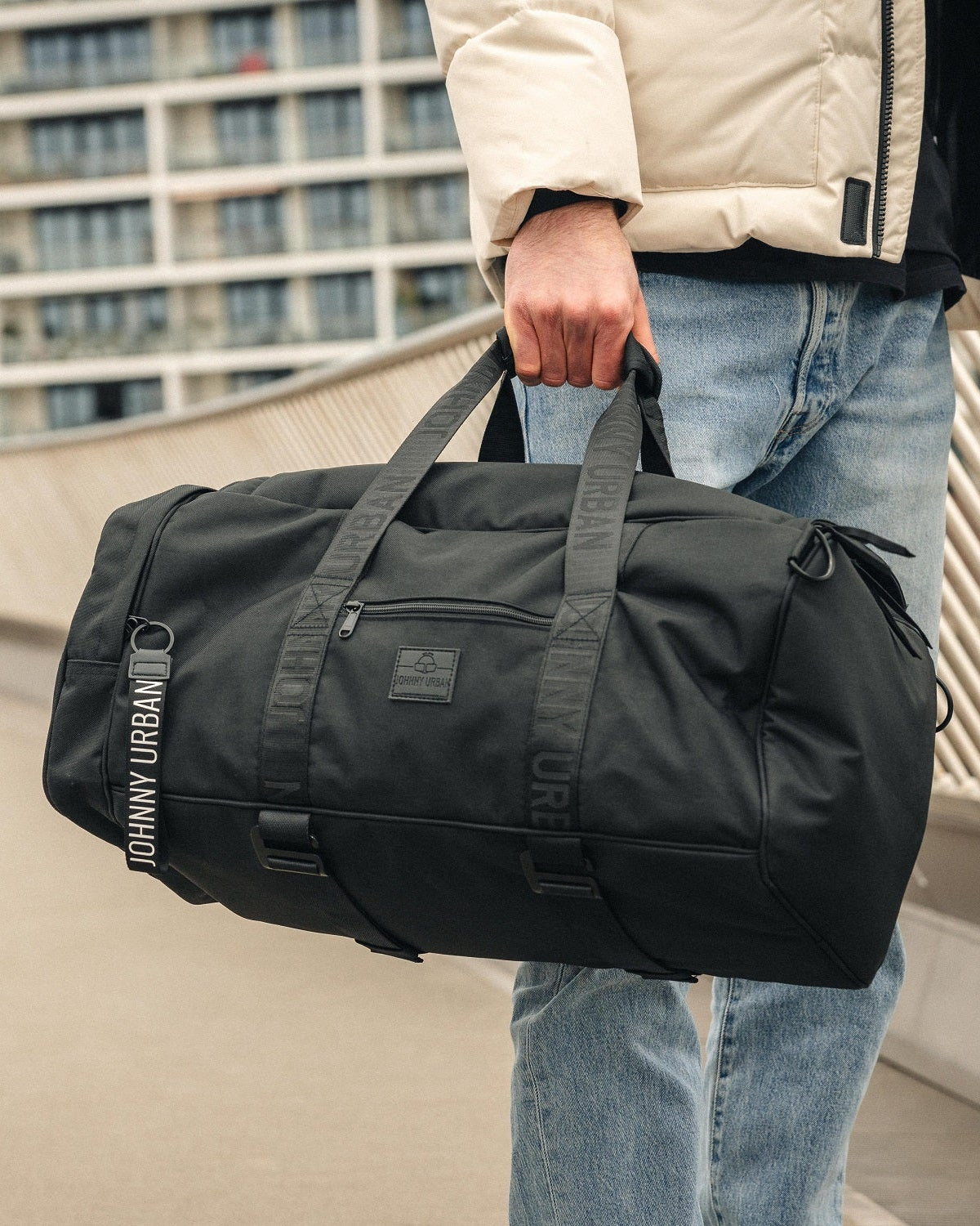 Schwarze Johnny Urban Duffle Bag 'Colin' aus robustem Material, getragen von Mann in beiger Jacke und Jeans
