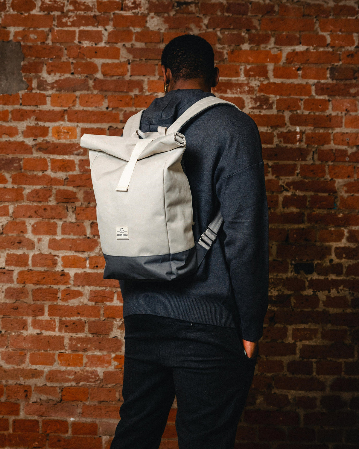 Mann trägt Rolltop-Rucksack 'Ryan' in Beige mit dunkelgrauem Boden vor Backsteinwand