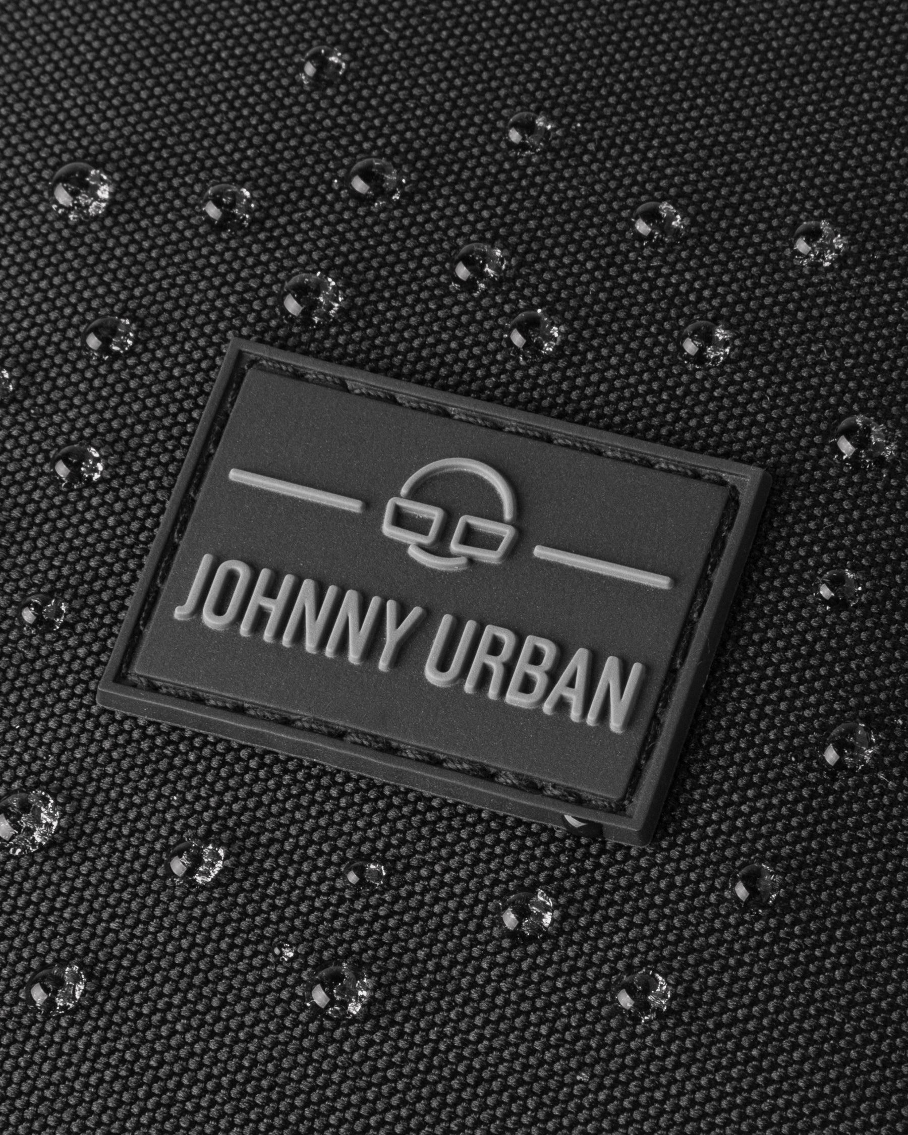 Nahaufnahme des Logos von Johnny Urban auf dem wasserfesten Stoff des Rucksacks ‚Nico‘ mit Wassertropfen