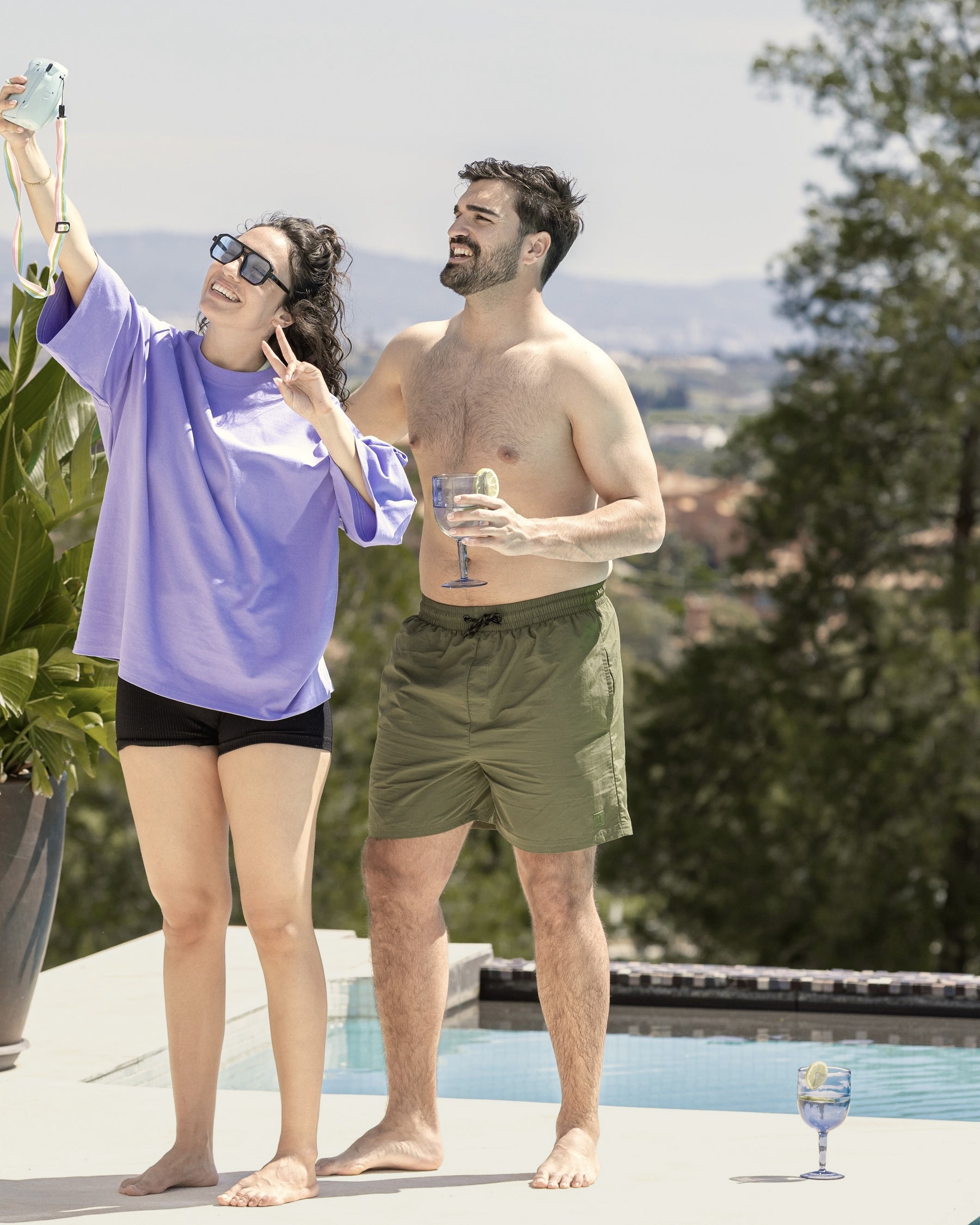 Mann in olivgrüner Badehose 'Jay' steht am Pool neben Frau in lila Oberteil und schwarzen Shorts, die Selfie macht