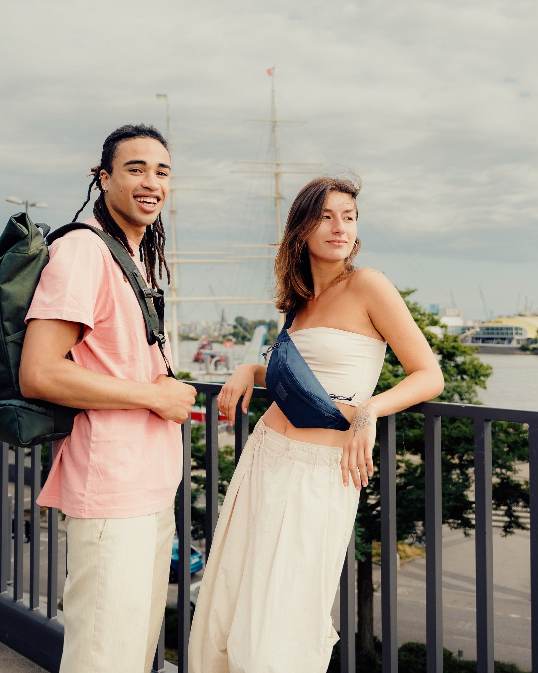 Junge Frau trägt die blaue Bauchtasche 'Erik Small' von Johnny Urban über dem Oberkörper, neben ihr steht ein junger Mann mit grünem Rucksack in urbaner Umgebung
