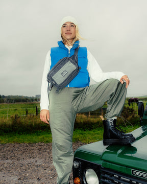 Frau trägt graue Bauchtasche 'Travis' von Johnny Urban, blaue Weste, weiße Mütze und schwarze Stiefel, steht mit Fuß auf grünem Defender