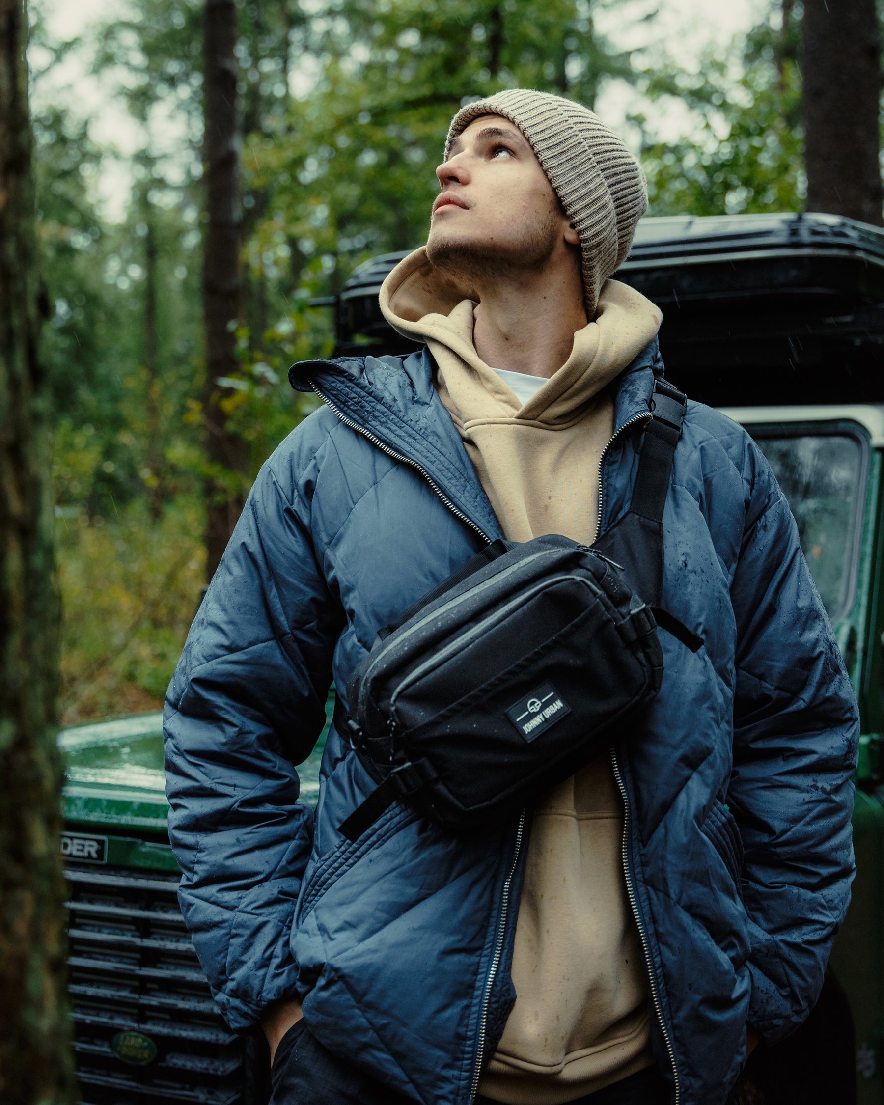 Mann trägt schwarze Johnny Urban Bauchtasche 'Travis' quer über die Brust, gesteppten blauen Mantel und beigefarbenen Hoodie, Waldhintergrund