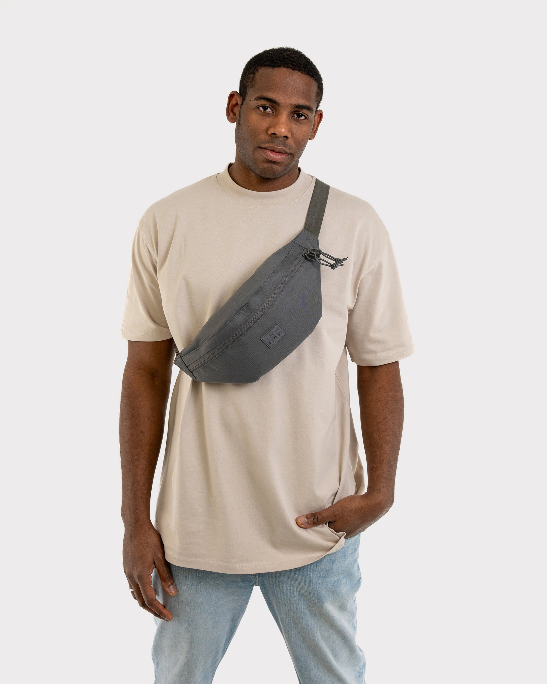 Mann trägt dunkelgraue Bauchtasche 'Erik Large' von Johnny Urban diagonal über beige T-Shirt und Jeans