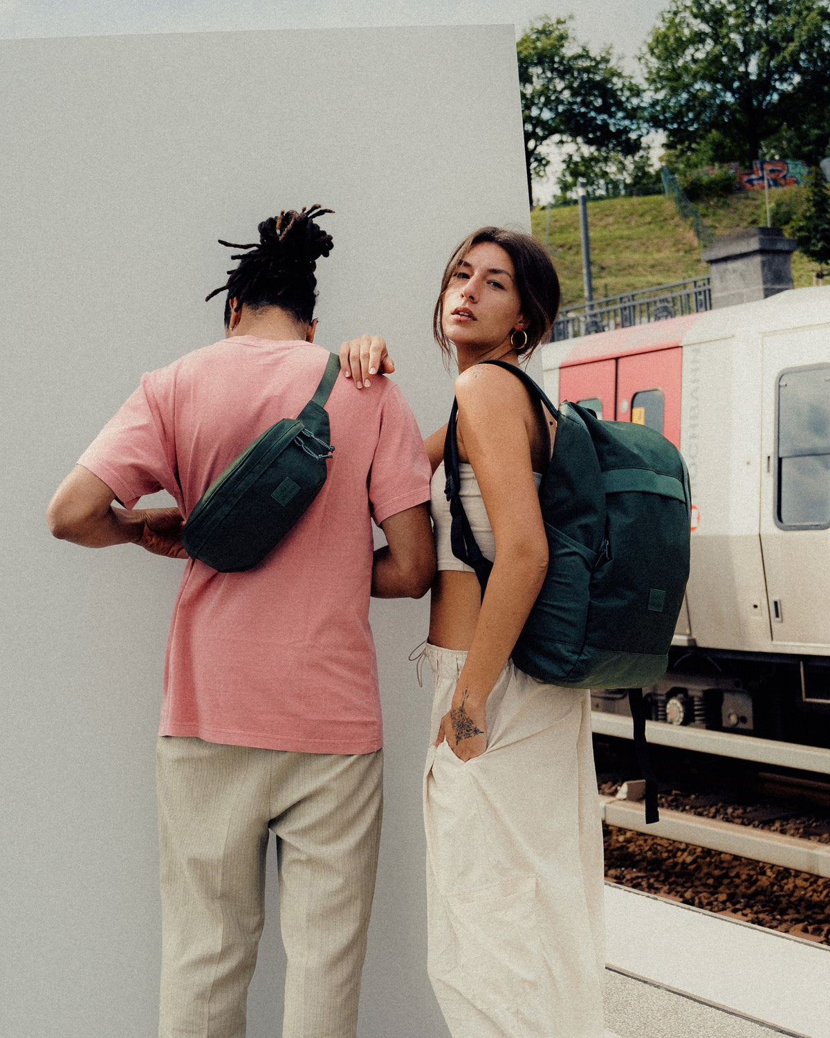 Mann trägt grüne Johnny Urban Bauchtasche 'Tom' quer über dem Rücken mit rosa T-Shirt und Frau mit grünem Rucksack neben Zug