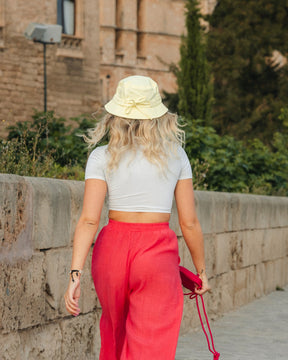 Frau von hinten mit gelbem Bucket Hat 'Gill', weißem Crop-Top und roten Hosen, die an einer Steinmauer vorbeigeht