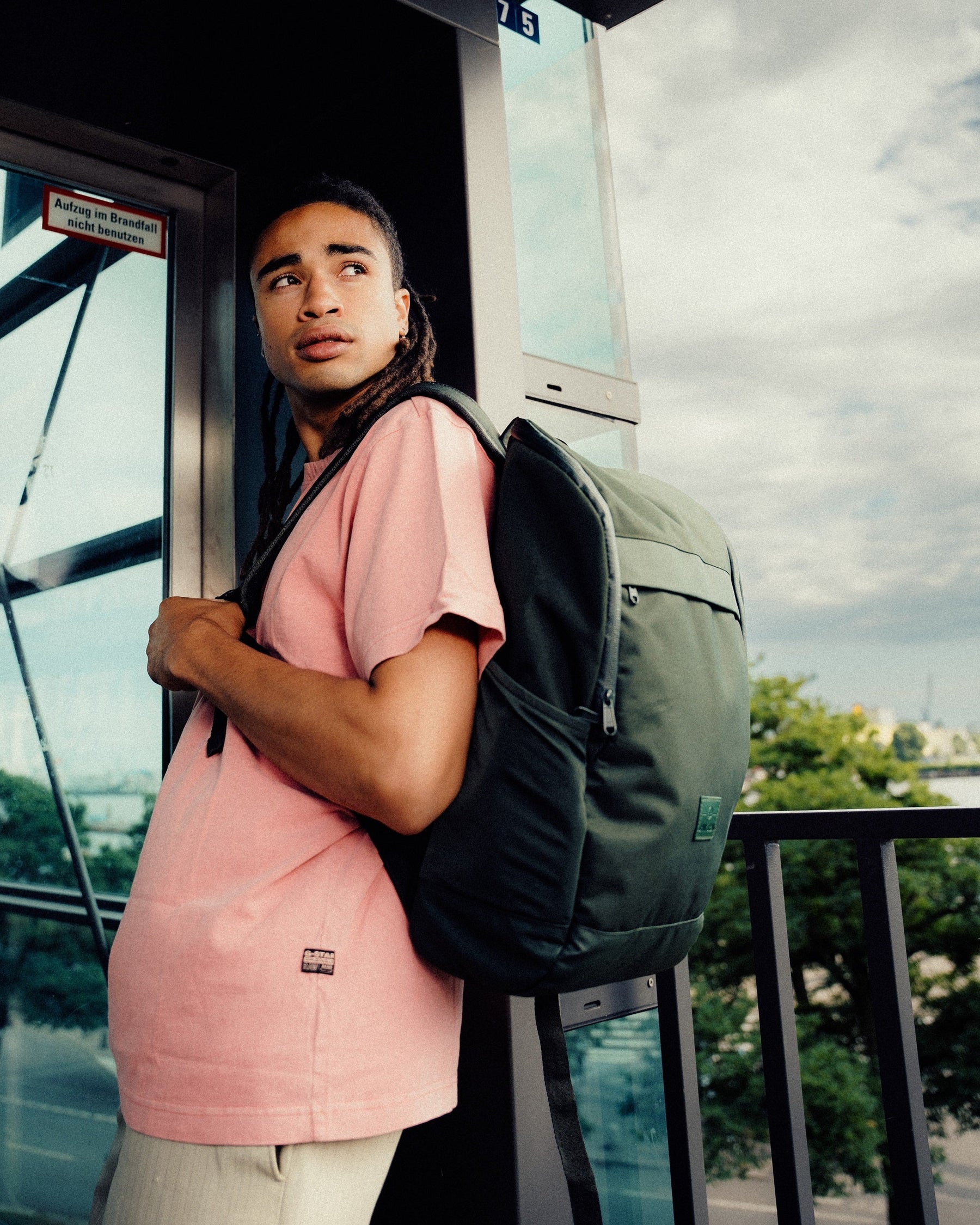 Mann mit Business-Rucksack 'Jasper' von Johnny Urban in Olivgrün und rosa T-Shirt stehend an einer Glasscheibe im urbanen Umfeld