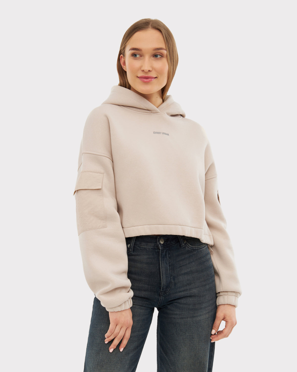 Frau trägt hellbeige Cropped Hoodie 'Carly' mit Kapuze und Taschen auf den Ärmeln, kombiniert mit dunklen Jeans