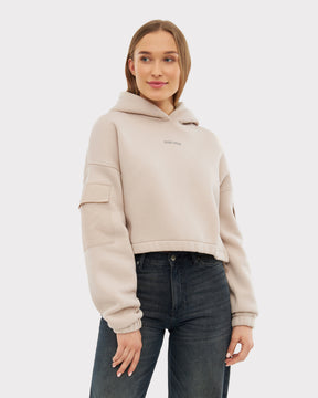 Frau trägt hellbeige Cropped Hoodie 'Carly' mit Kapuze und Taschen auf den Ärmeln, kombiniert mit dunklen Jeans