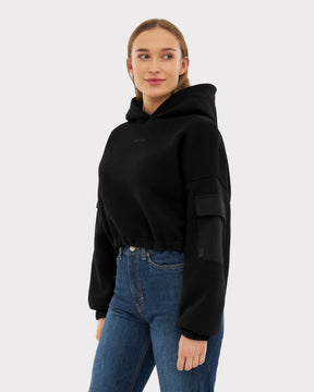 Frau trägt schwarzen Cropped Hoodie 'Carly' mit Kapuze und Tasche am Ärmel, kombiniert mit blauen Jeans