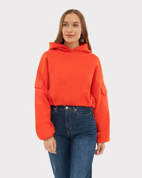 Junge Frau trägt rotes, kurzes Hoodie 'Carly' mit Taschen an den Ärmeln und blauen Jeans