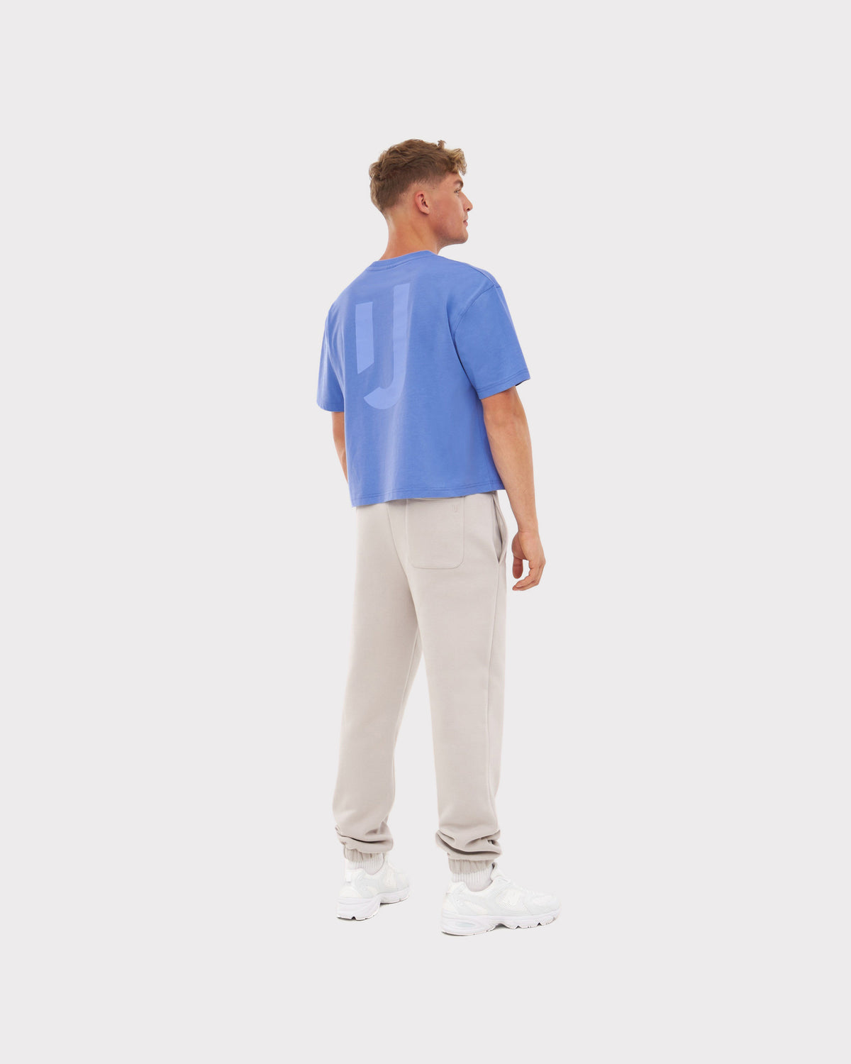 Mann trägt blaues, oversize Cropped T-Shirt 'Zoey JU' mit kurzem Ärmel und Logo auf dem Rücken, beige Jogginghose und weiße Sneaker