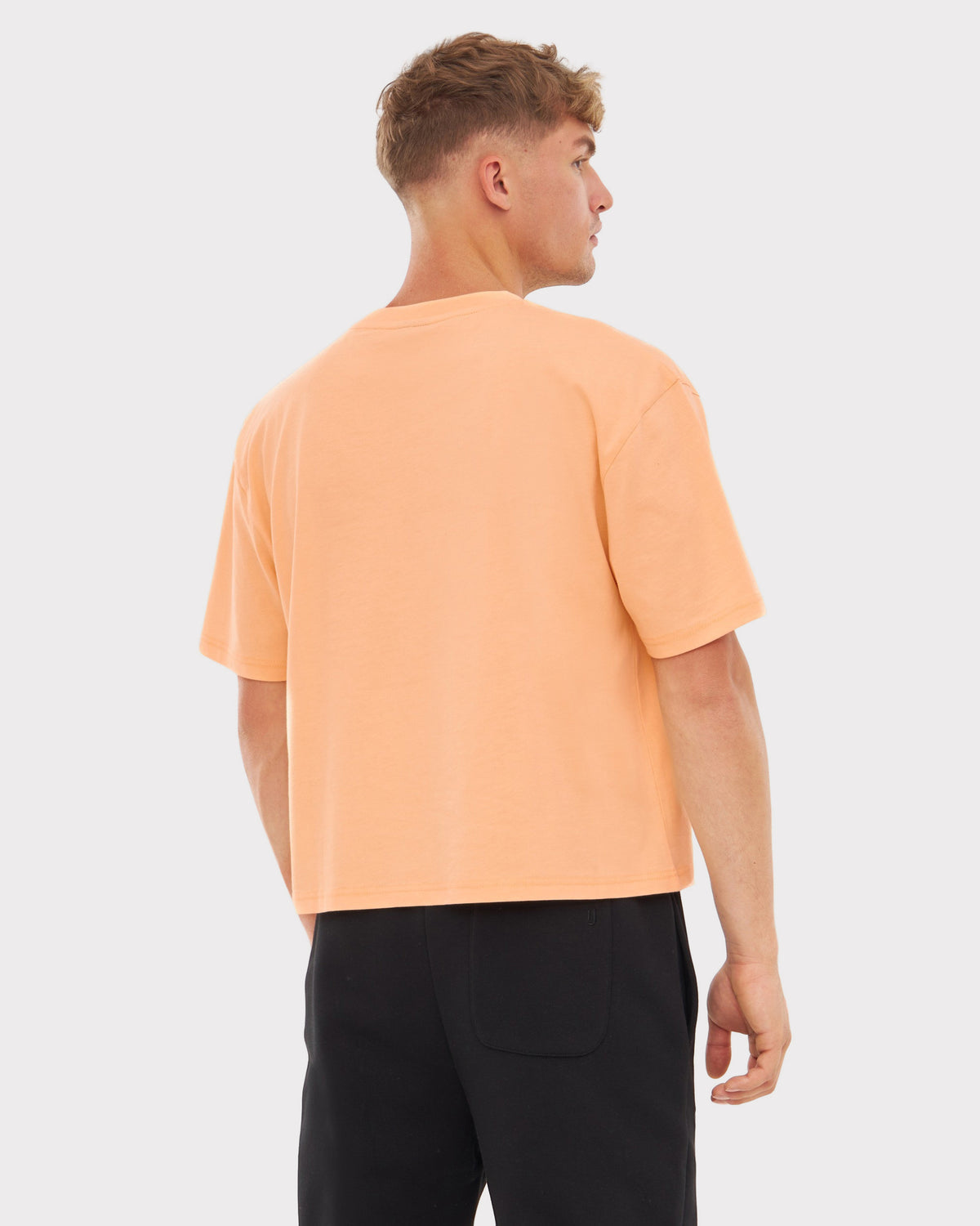 Mann trägt 'Zoey' Oversize Cropped T-Shirt in Pfirsich, Rückansicht, kombiniert mit schwarzen Hosen