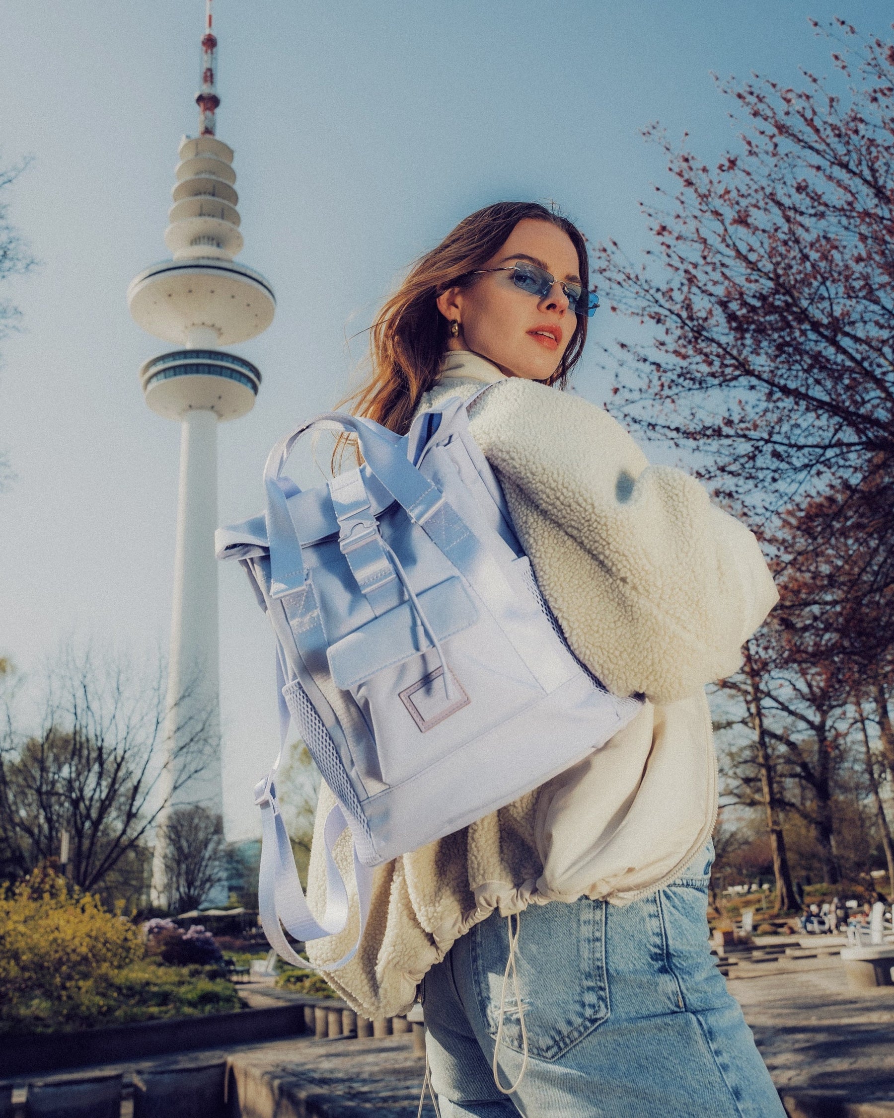 Frau mit lila Damenrucksack 'Jules' trägt weiße Jacke und Jeans vor Fernsehturm im Park