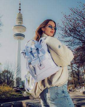 Frau mit lila Damenrucksack 'Jules' trägt weiße Jacke und Jeans vor Fernsehturm im Park