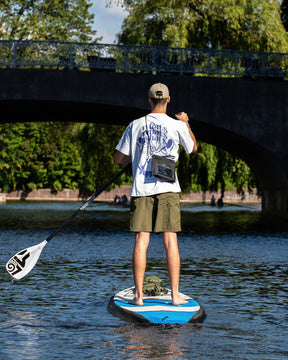 Rückansicht eines Mannes auf einem Stand-up-Paddle-Board mit Dry Bag 'Alfie' am Körper, umgeben von Wasser und Brücke im Hintergrund