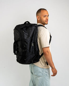 Mann trägt schwarzen Duffle Bag 'Shawn Large' auf Rücken, beige T-Shirt, helle Jeans
