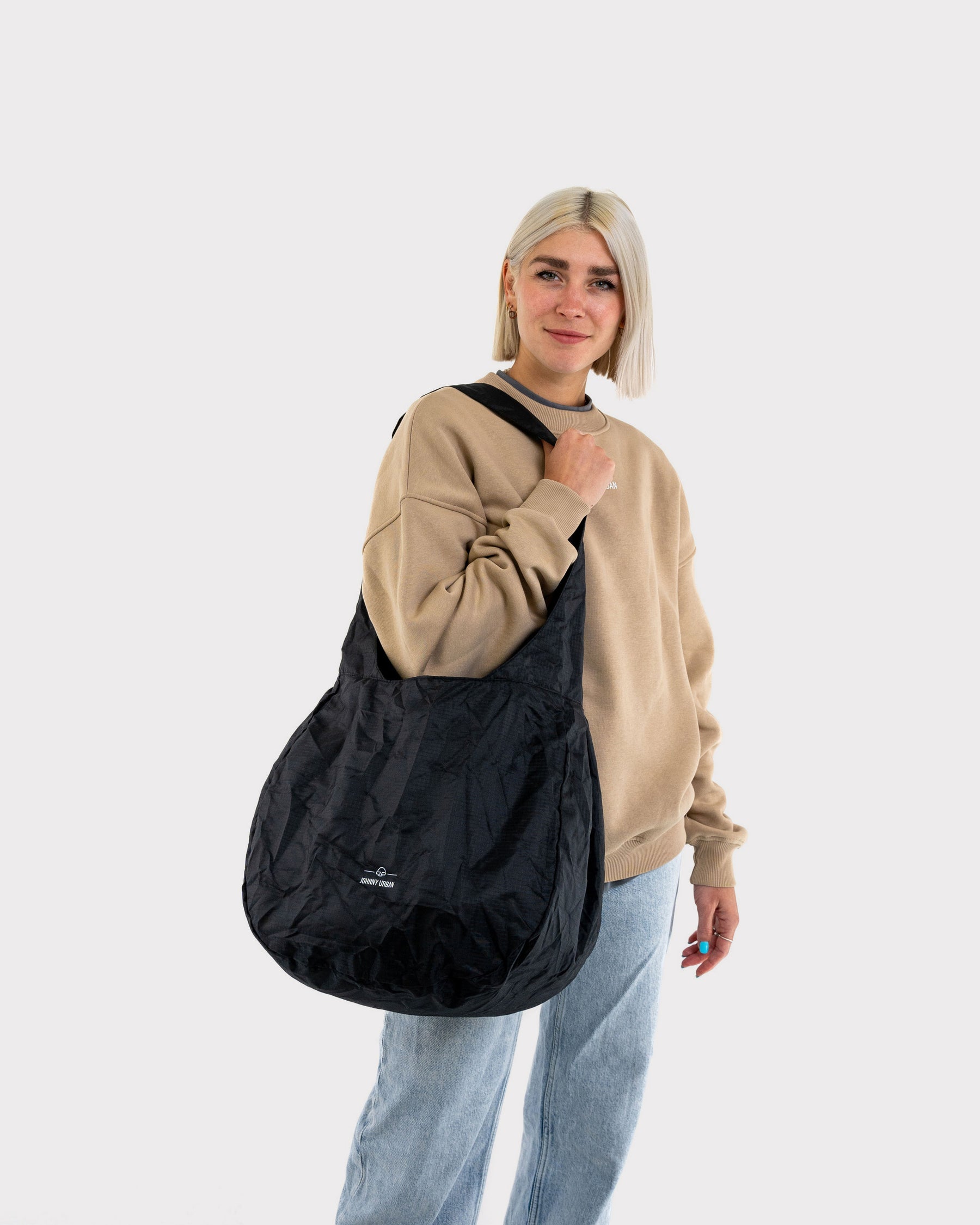 Frau trägt beige Sweatshirt und hellblaue Jeans mit großer, schwarzer faltbarer Einkaufstasche 'Lee' von Johnny Urban