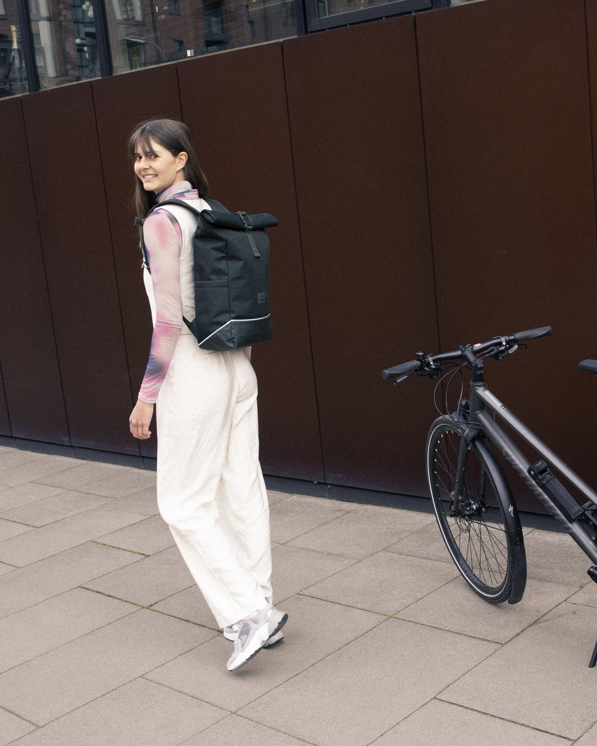 Frau trägt dunkelgrünen Rolltop Rucksack 'Robin Medium' von Johnny Urban neben Fahrrad auf urbanem Gehweg