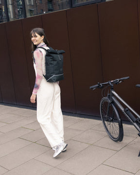 Frau trägt dunkelgrünen Rolltop Rucksack 'Robin Medium' von Johnny Urban neben Fahrrad auf urbanem Gehweg