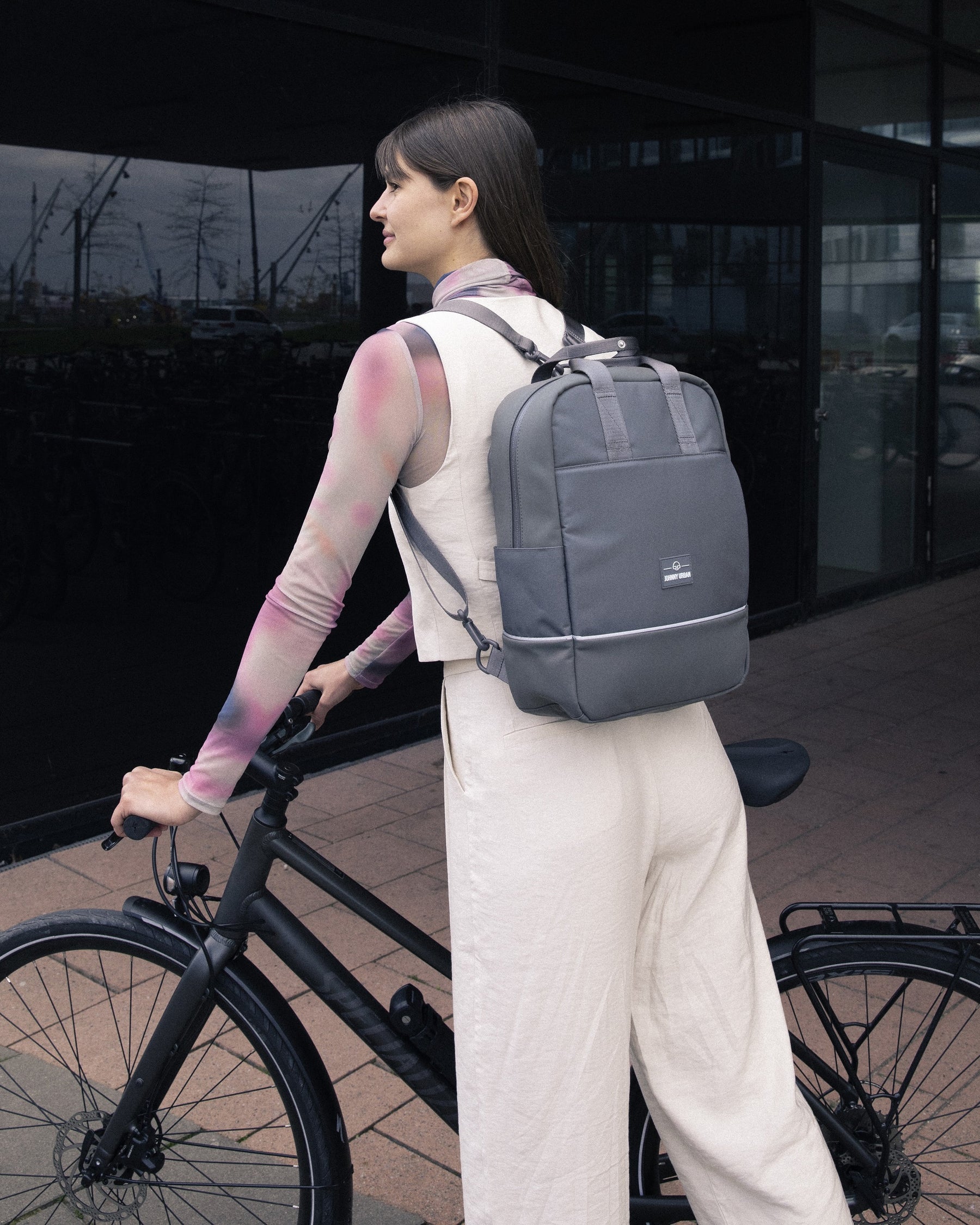 Frau mit grauem Johnny Urban Rucksack 'Jona Medium' lehnt an schwarzem Fahrrad