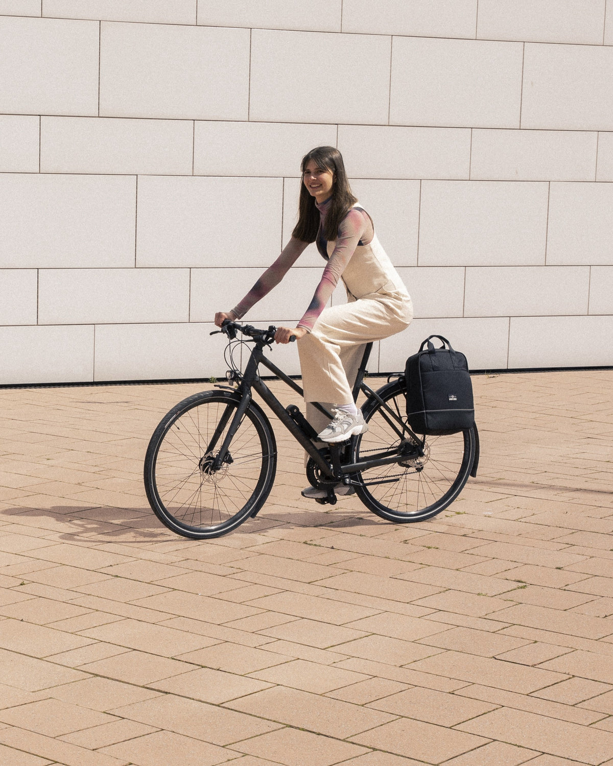 Frau mit langem braunem Haar fährt ein schwarzes Fahrrad mit beigem Outfit, daneben hängt der schwarze Rucksack 'Jona Medium' von Johnny Urban am Gepäckträger