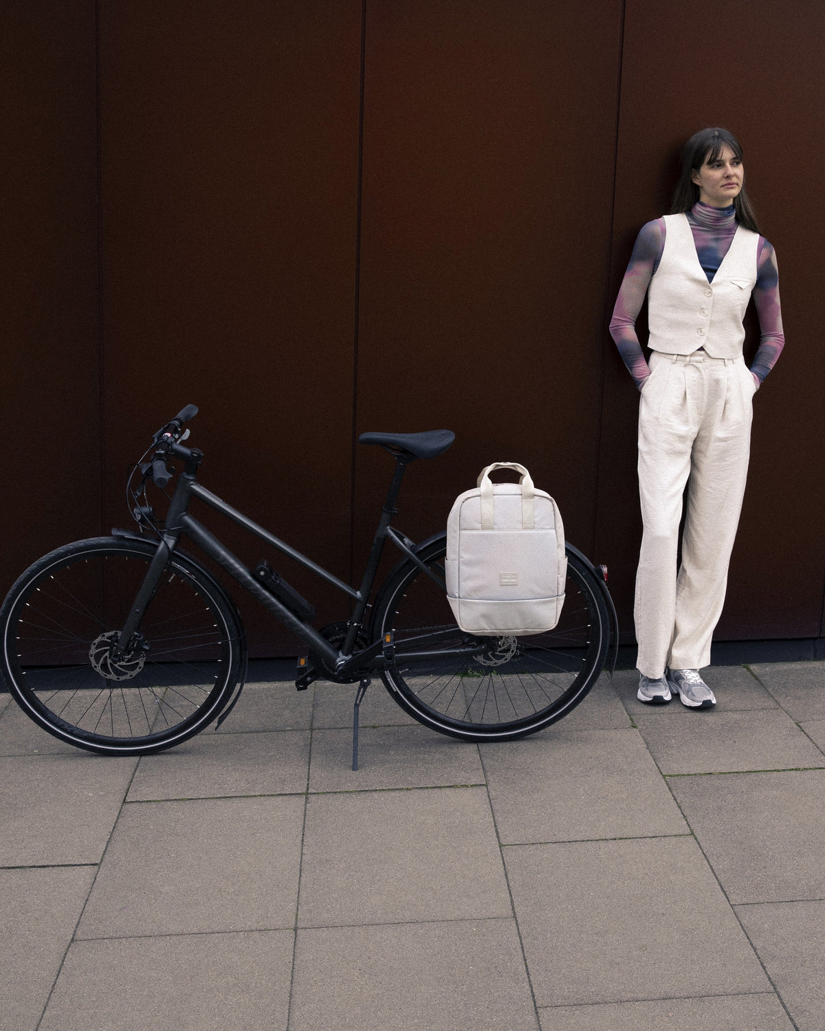 Fahrrad mit beigefarbenem Johnny Urban Rucksack 'Jona Medium' am Gepäckträger, daneben steht eine Frau in heller Hose und Weste