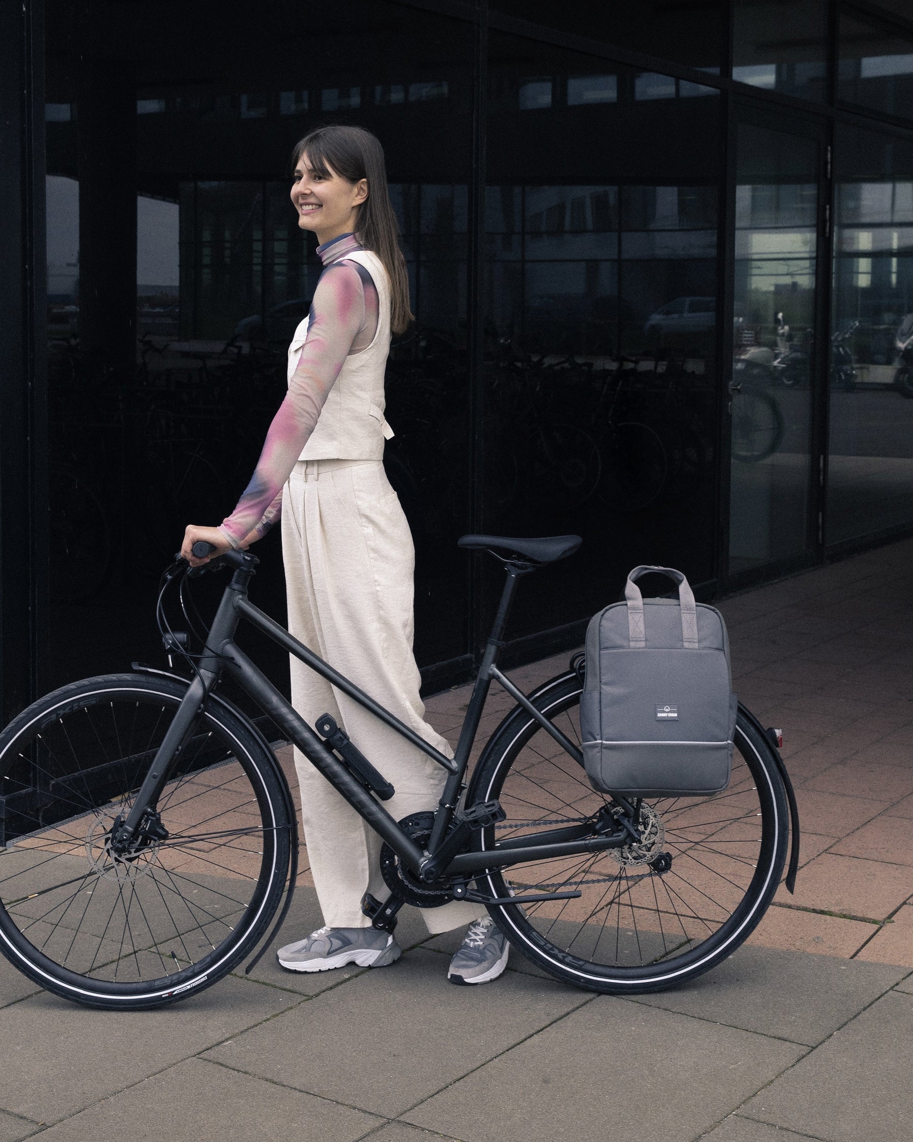 Frau steht neben Fahrrad mit grauem Rucksack 'Jona Medium' am Gepäckträger