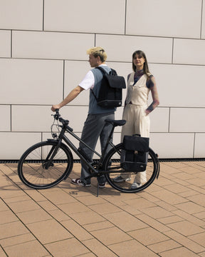 Mann mit schwarzem Rucksack 'Mika' auf einem schwarzen Fahrrad, Frau neben ihm mit schwarzem Rucksack 'Mika' neben Fahrrad