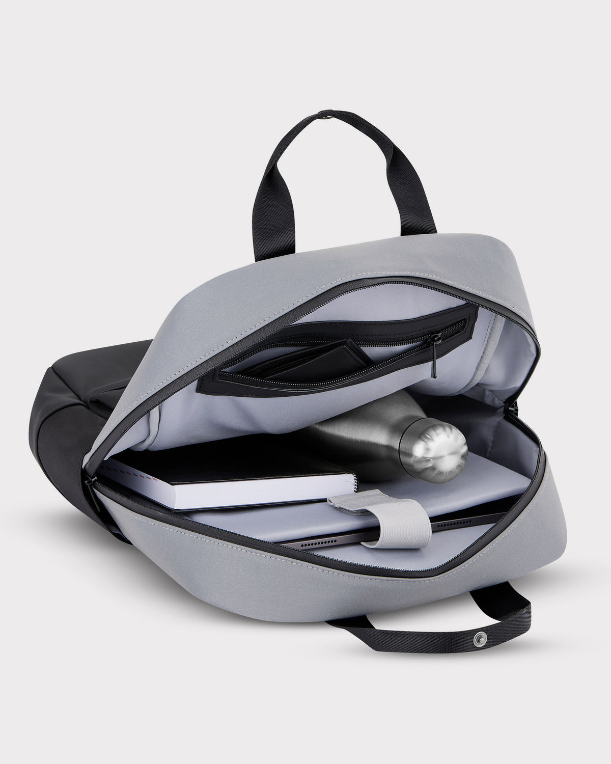 Geöffneter grauer Rucksack 'Jona Medium' mit reflektierendem Material, enthält Wasserflasche, Notizbuch und Tablet