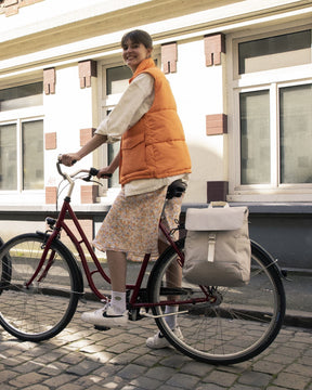Frau in orangefarbener Weste sitzt auf Fahrrad mit beigefarbenem Rucksack 'Mika' am Gepäckträger
