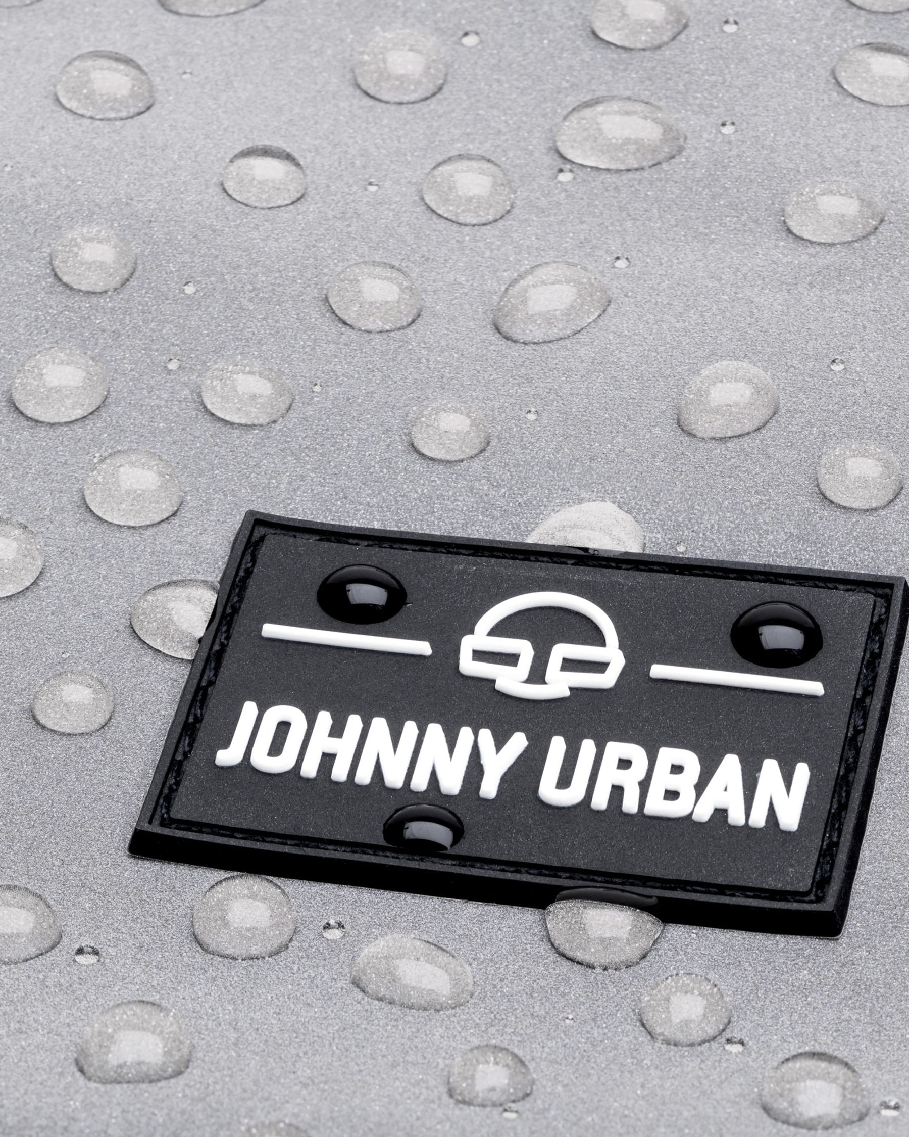 Nahaufnahme eines schwarzen Johnny Urban Logos auf silbernem, wasserabweisendem Material mit Wassertropfen