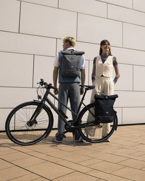 Mann mit grauem Rolltop Rucksack 'Robin Medium' von Johnny Urban, Frau steht daneben, schwarzes Fahrrad im Vordergrund
