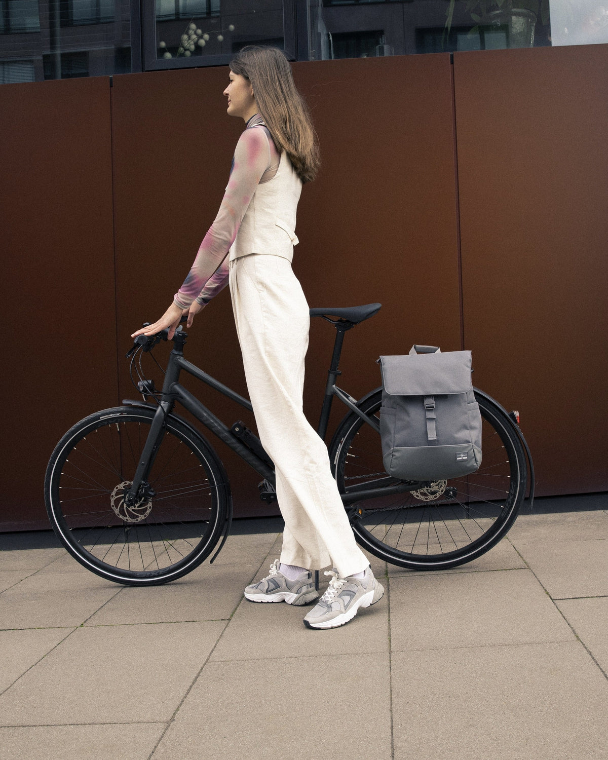 Frau in heller Kleidung steht neben schwarzem Fahrrad mit grauer Fahrradtasche Rucksack 'Mika' Bike am Gepäckträger
