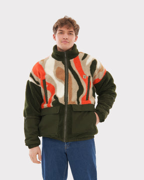 Mann trägt Teddy Fleecejacke 'Carson' mit olivgrünen, weißen, braunen und orangen Mustern und blauer Jeans