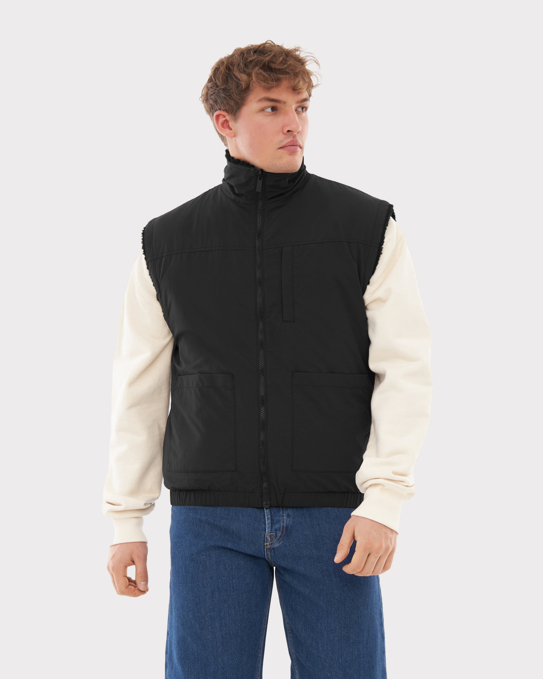Mann trägt schwarze, moderne, wendbare Teddy Fleeceweste 'Bryson' über cremefarbenem Pullover und blauen Jeans