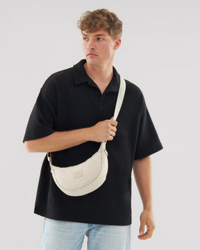 Mann trägt beige Half Moon Bag 'Kaia Small' über der Schulter, trägt schwarzen Poloshirt und helle Jeans