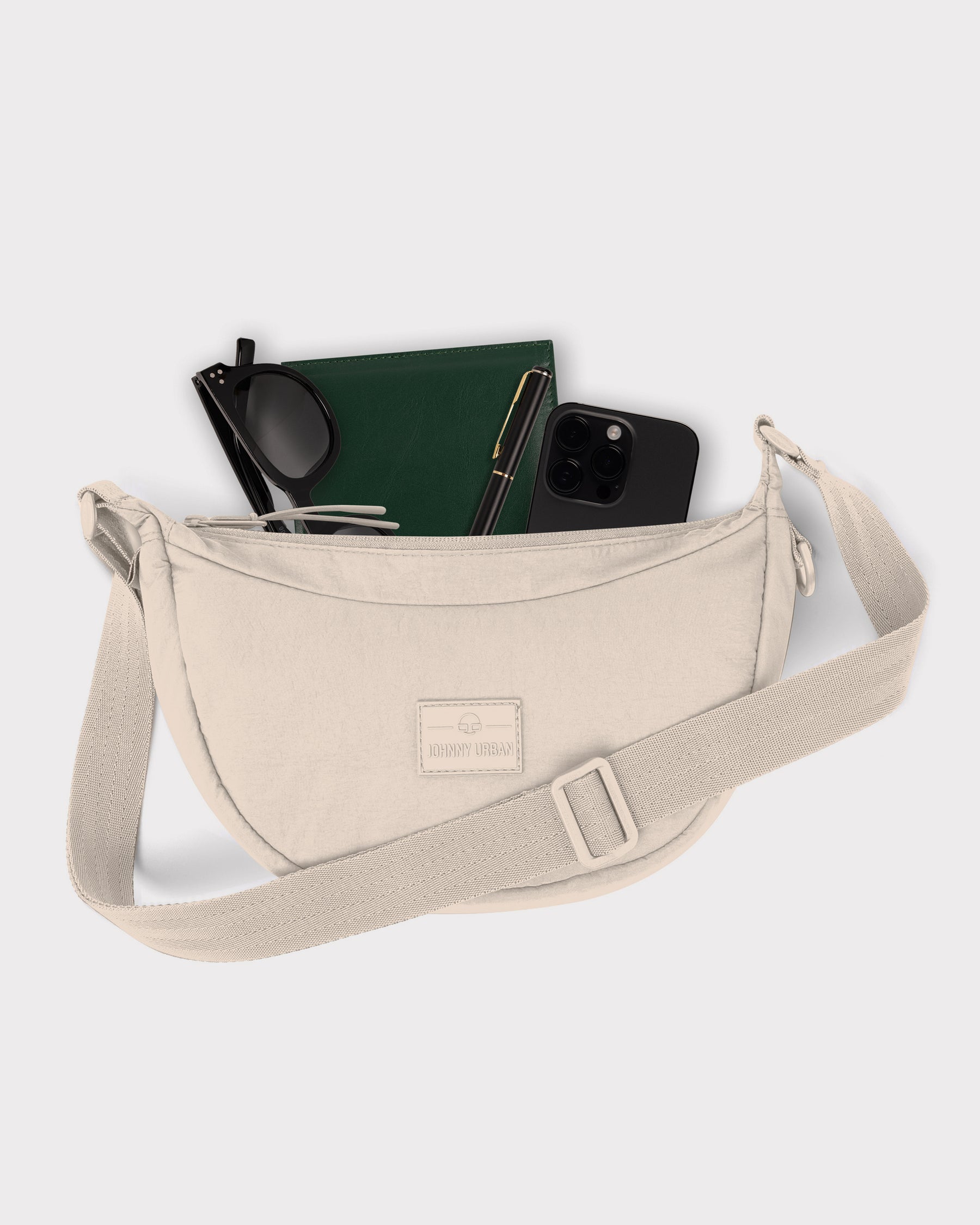 Beige kleine Half Moon Bag 'Kaia Small' von Johnny Urban mit Sonnenbrille, grünem Notizbuch, Stift und schwarzem Smartphone