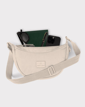 Beige kleine Half Moon Bag 'Kaia Small' von Johnny Urban mit Sonnenbrille, grünem Notizbuch, Stift und schwarzem Smartphone
