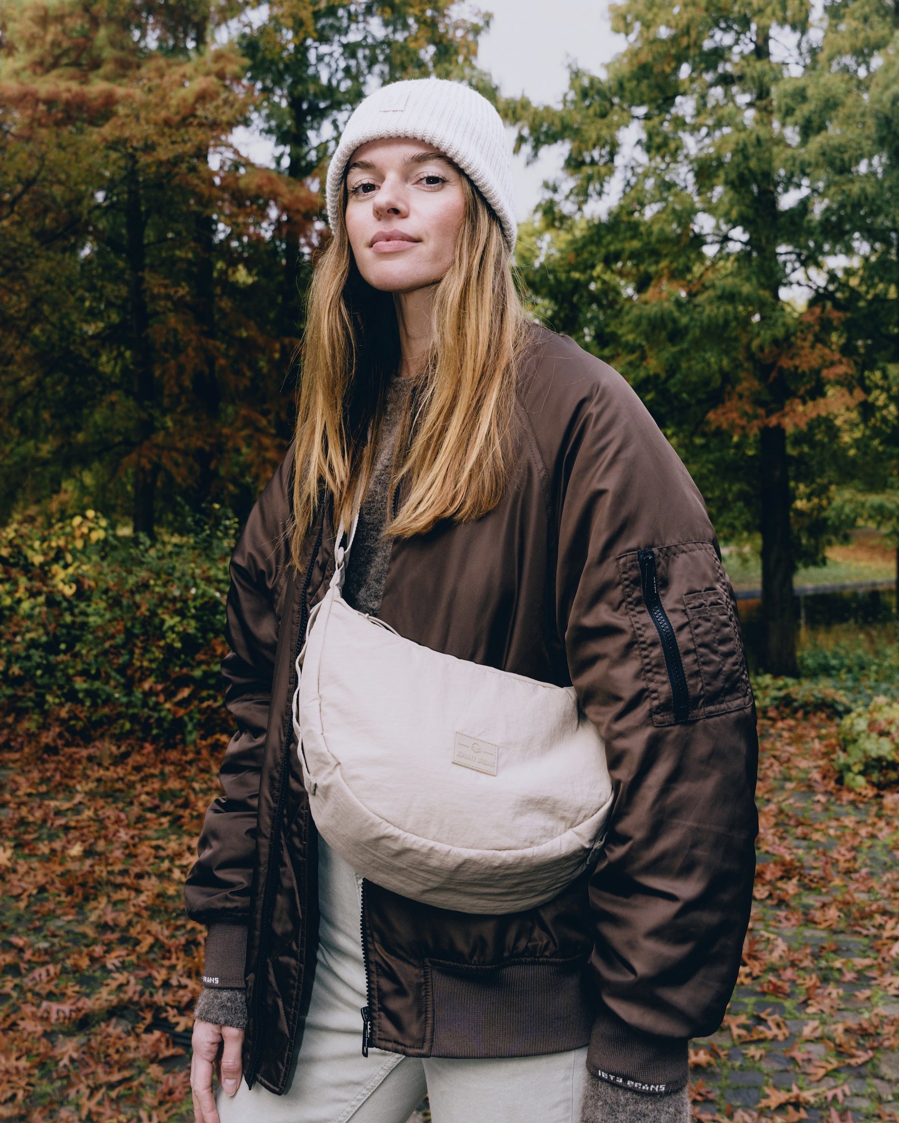Frau mit beige gepolsterter Half Moon Bag 'Kaia Medium', brauner Jacke und weißer Mütze im herbstlichen Park