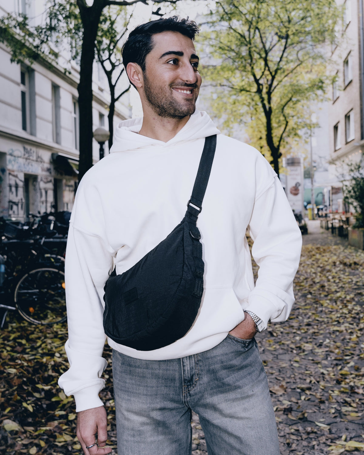 Mann mit schwarzer Half Moon Bag 'Kaia Medium', weißem Hoodie und blauen Jeans auf herbstlicher Straße