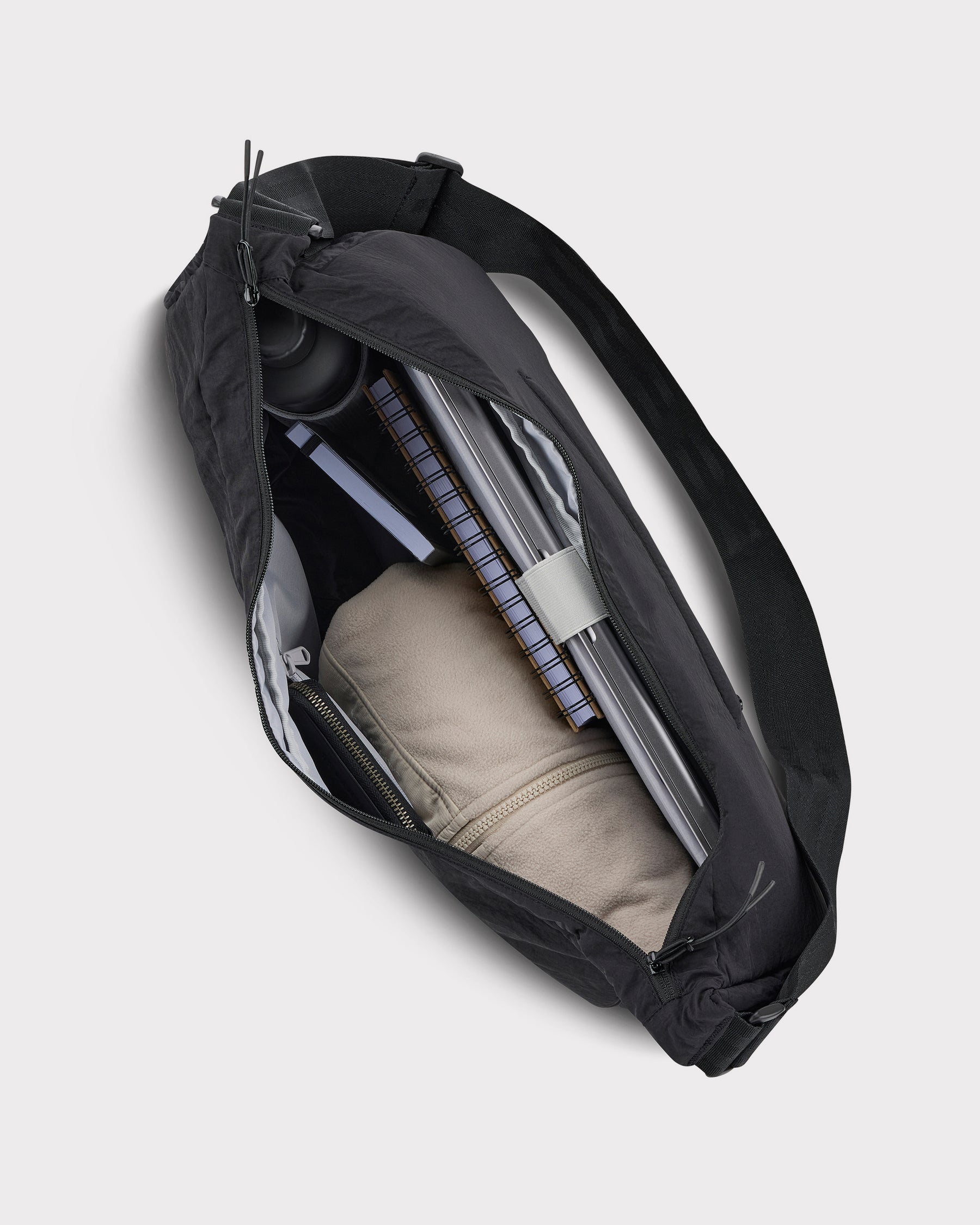 Schwarze Half Moon Bag 'Kaia XL' mit Reißverschluss, gefüllt mit Laptop, Notizbuch, Trinkflasche und beige Jacke